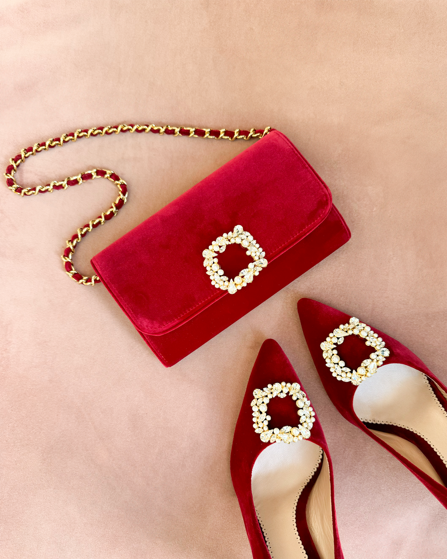 Red suede heels with a pointed toe and embellished buckle detail are positioned on a soft, neutral background. A matching red suede clutch with a decorative buckle sits beside the shoes, creating a coordinated look. The image is taken from a top-down angle, showcasing the elegant design of both the heels and the bag. image
