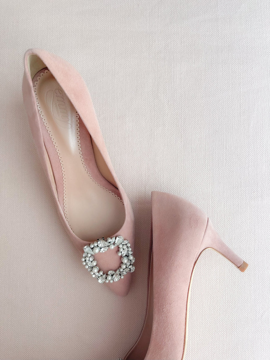 Pink suede heels featuring a decorative rhinestone buckle on the vamp. The shoes are positioned on a soft, light-colored fabric background, showcasing a side view of the heel. The angle highlights the elegant silhouette of the shoes. image