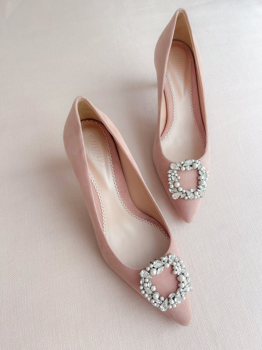 Pink suede heels featuring a decorative crystal buckle on the toe. The shoes are displayed on a soft, light-colored background, showcasing their elegant design. The angle captures both shoes from above, highlighting the pointed toe and the intricate embellishment. image