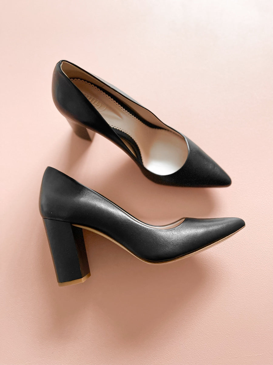 Black leather heels with a block heel, approximately 3 inches in height. The shoes are displayed on a soft pink background, showcasing a top-down view. The pointed toe design adds a sleek touch, and the interior is visible, indicating a polished finish. image
