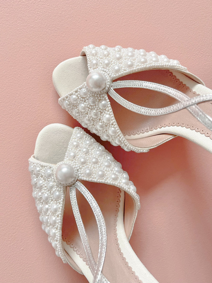 Elegant white heels adorned with pearl embellishments, featuring a delicate crisscross strap design. The shoes are positioned against a soft pink background, showcasing their intricate detailing. The heel height is approximately 3 inches. image