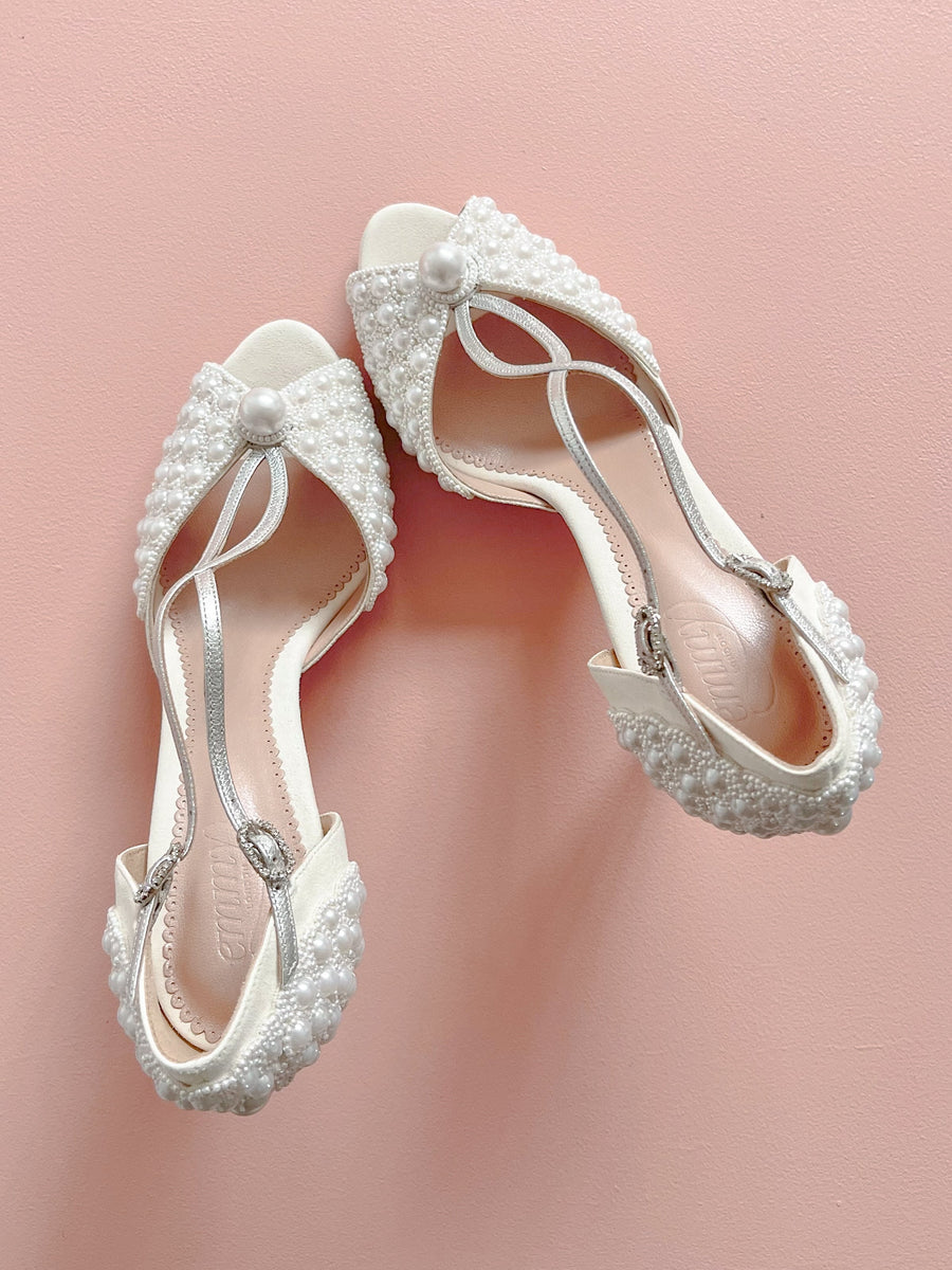 Elegant white shoes adorned with intricate pearl detailing and a delicate strap design. The shoes are positioned on a soft pink background, showcasing their unique texture and embellishments. The angle captures the shoes from above, highlighting their stylish silhouette. image