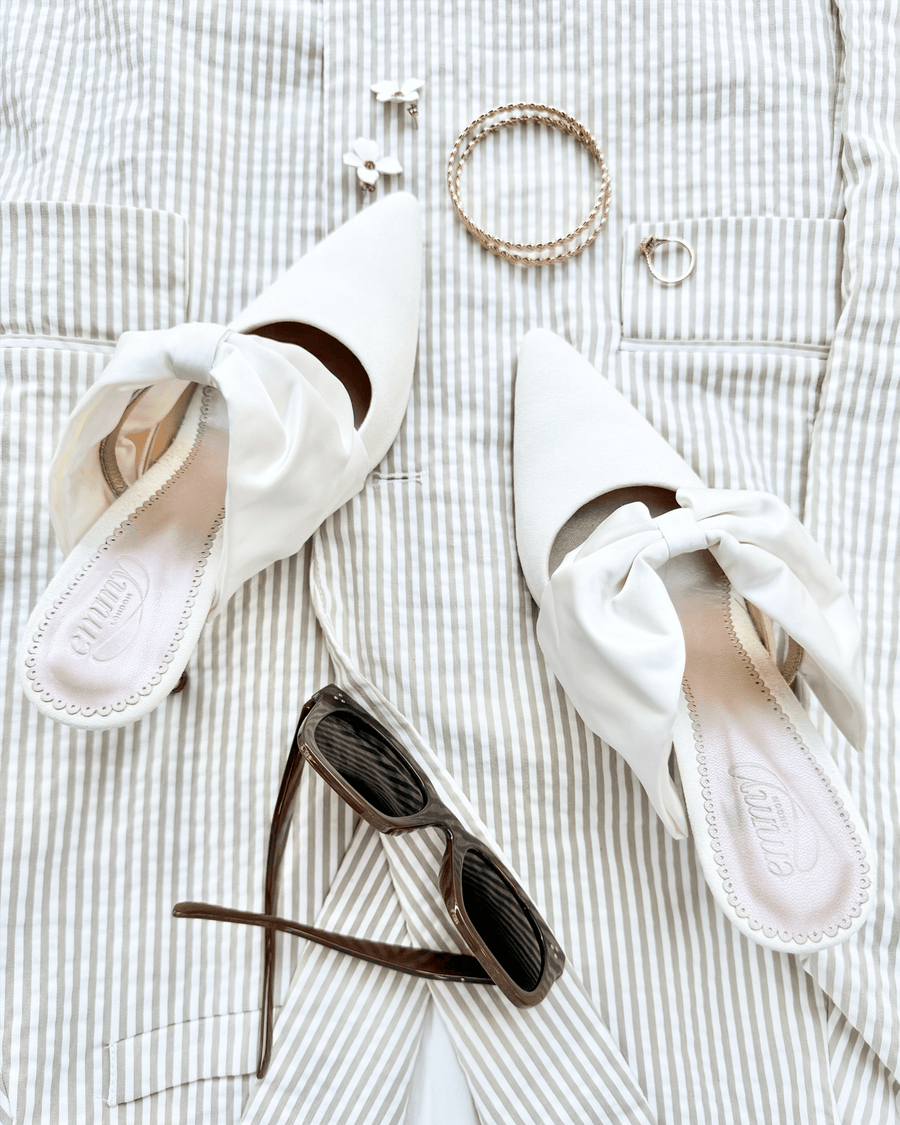 White pointed-toe mules with a bow detail on the front, placed on a light striped fabric background. The shoes are made of a smooth material and have a moderate heel height. Accompanying accessories include a pair of sunglasses, a bracelet, and earrings, arranged neatly beside the shoes. image