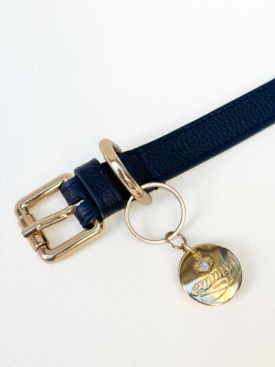 A close-up image of a navy blue leather belt featuring a shiny gold buckle and a decorative charm. The charm is circular with a diamond accent and the word "Mantra" inscribed on it. The background is plain white, highlighting the details of the belt and charm. image