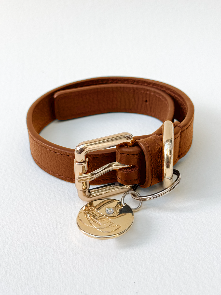 Brown leather bracelet with a gold buckle and charm, displayed against a white background. The bracelet is flat and not worn by anyone in the image. image