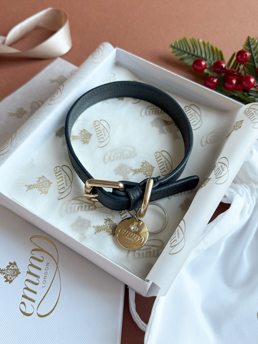 Black leather bracelet with a gold buckle and charm, presented in a white box with the "Emmy London" logo. The background features a warm brown surface and decorative greenery with red berries, adding a festive touch. The image is taken from a top-down angle, showcasing the bracelet's details clearly. image