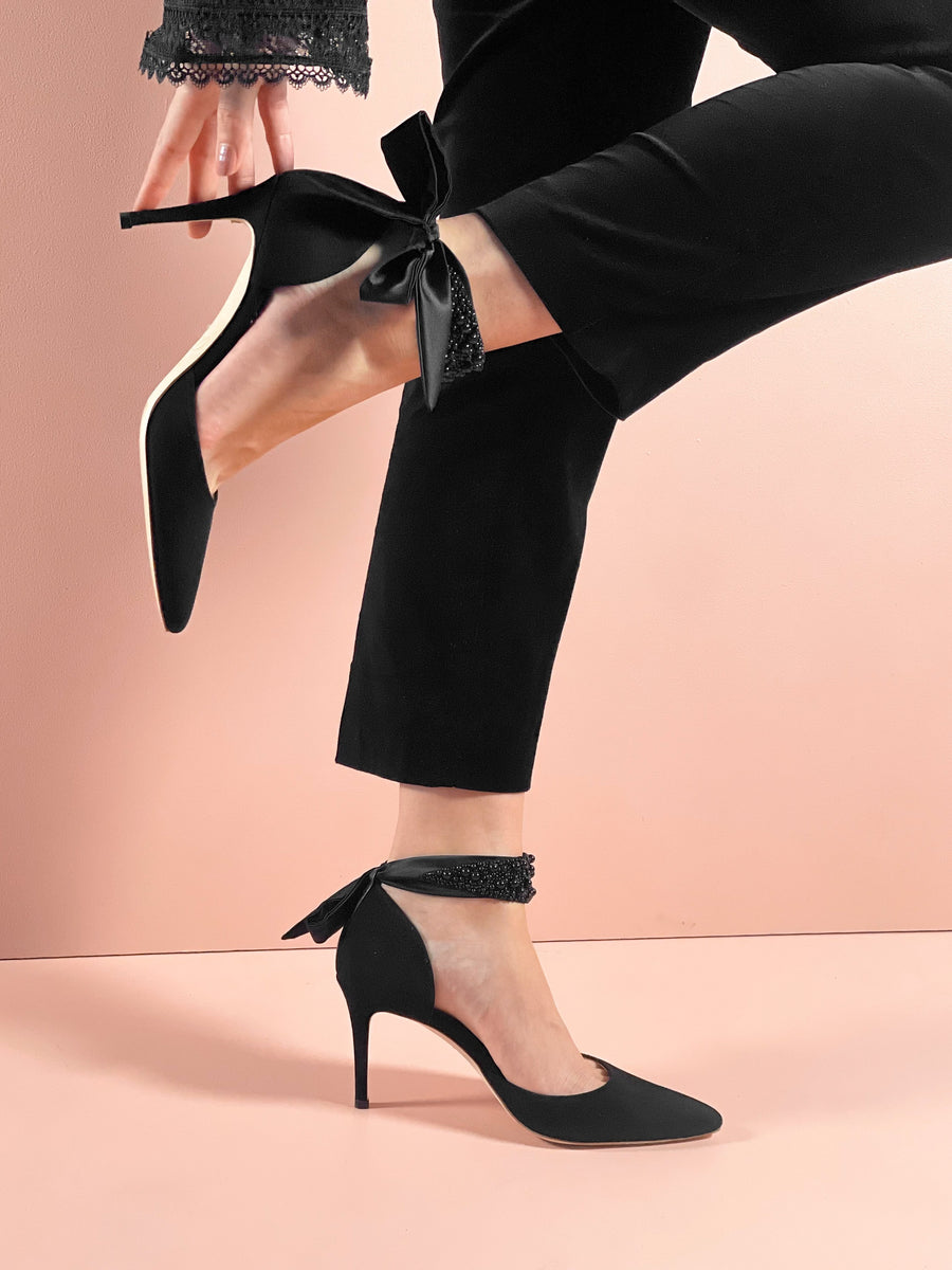 Black suede heels with a pointed toe and a delicate bow detail at the ankle. The model is wearing tailored black pants, and the image is taken from a side angle against a soft pink background, highlighting the elegant silhouette of the shoes. The heel height is approximately 10 cm. image