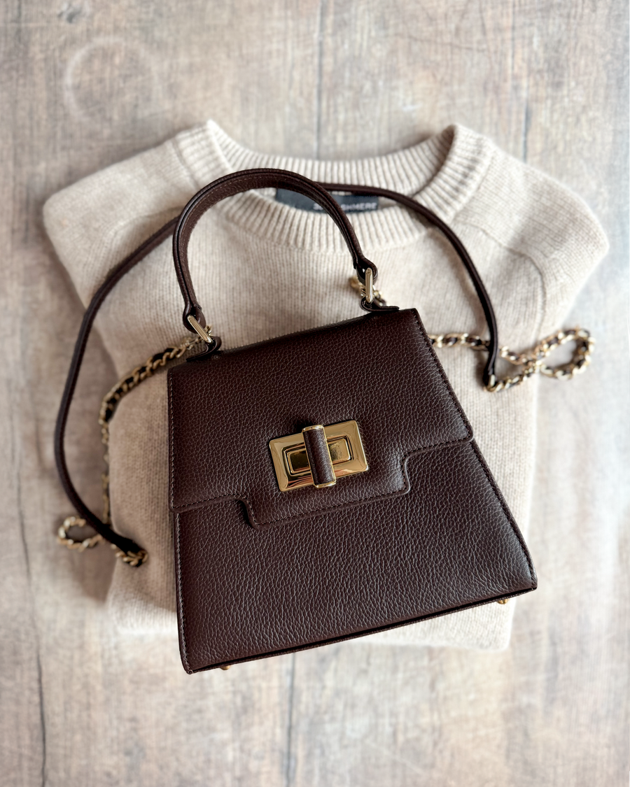 Brown leather handbag resting on a light beige sweater. The bag features a structured design with a gold-tone clasp and a detachable chain strap. The image is taken from a top-down angle, showcasing the texture of both the bag and the sweater. image