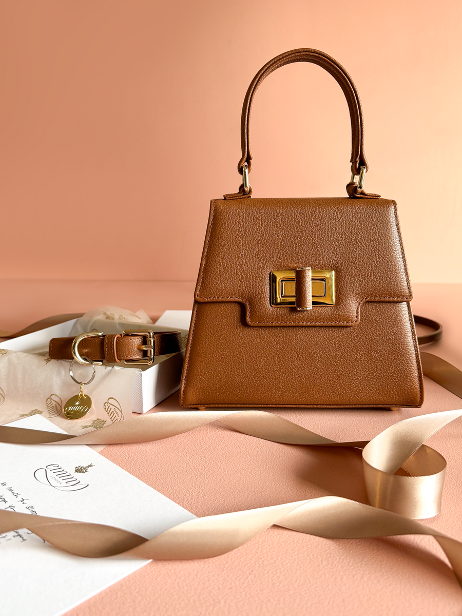 A structured brown handbag with a gold clasp sits elegantly against a soft peach background. The bag features a top handle and is accompanied by a matching belt, both resting on a textured surface. A delicate ribbon and a note with a subtle design are also present, adding a touch of sophistication to the composition. image
