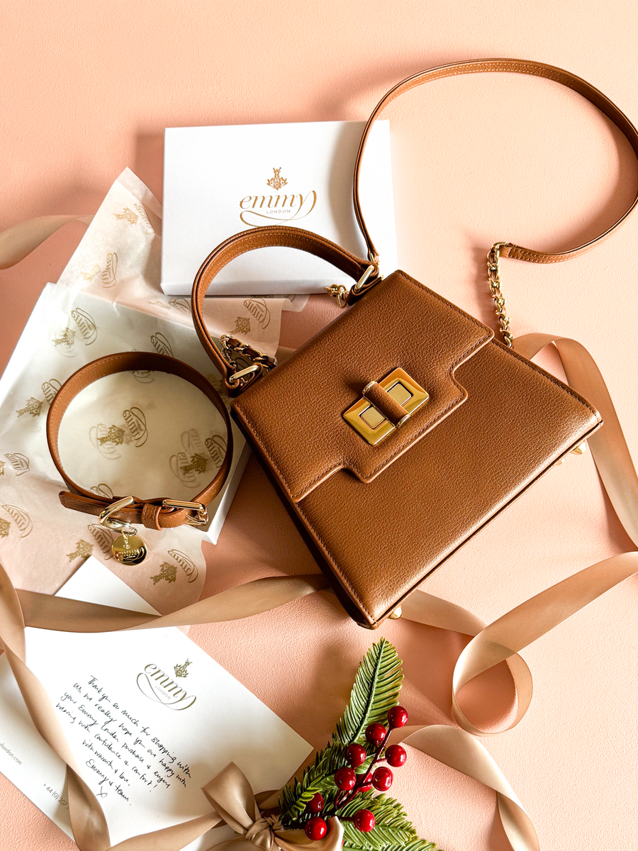 Brown leather handbag with a structured design, featuring a gold-tone clasp. The bag is placed on a soft pink background, accompanied by a gift box and decorative tissue paper. A matching strap is included, and the scene is adorned with festive greenery and red berries, creating a warm, inviting atmosphere. image