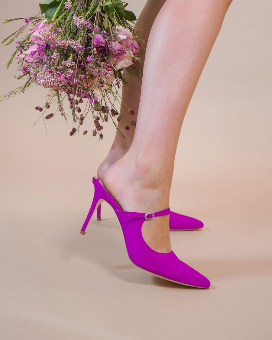 Bright pink high-heeled shoes with a pointed toe and a delicate ankle strap, showcased on a model's foot. The background is a soft beige, enhancing the vibrant color of the shoes. The model is holding a bouquet of flowers, adding a touch of elegance to the overall presentation. image