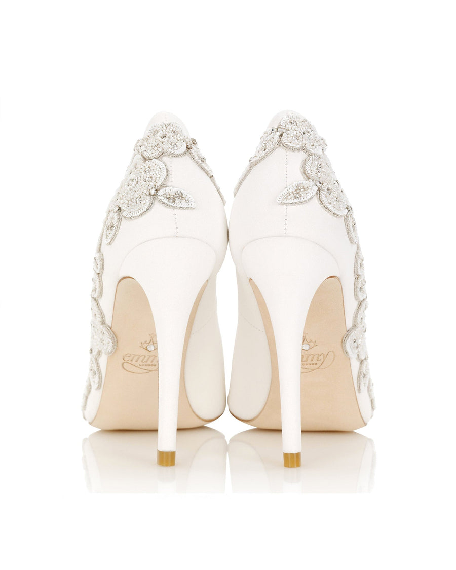 Elegant white heels adorned with intricate floral embellishments, featuring a sleek stiletto design. The image captures the back view of the shoes against a clean, white background, highlighting the detailed craftsmanship. The heels are high and slender, emphasizing a sophisticated silhouette. image