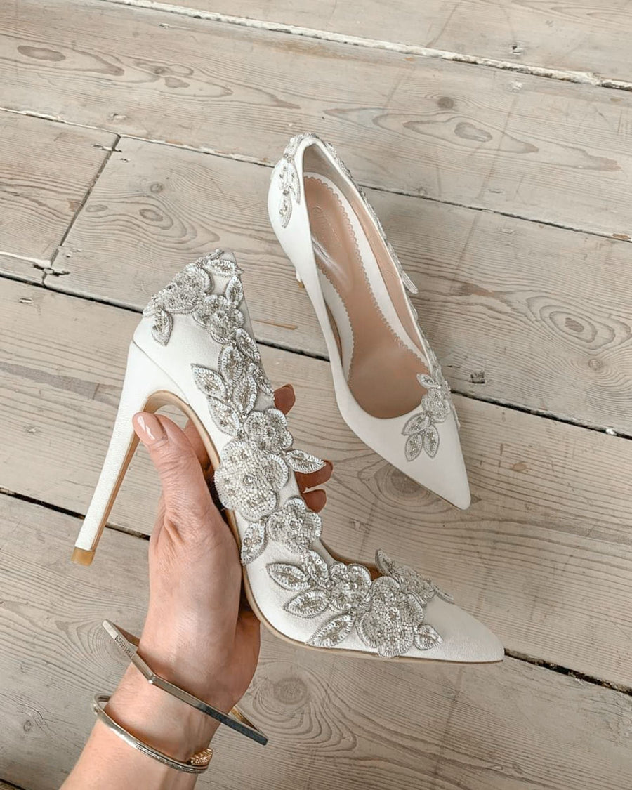 Elegant white heels adorned with intricate floral embellishments, held in a hand against a light wooden floor background. The shoes feature a pointed toe and a stiletto heel, showcasing a sophisticated design. image