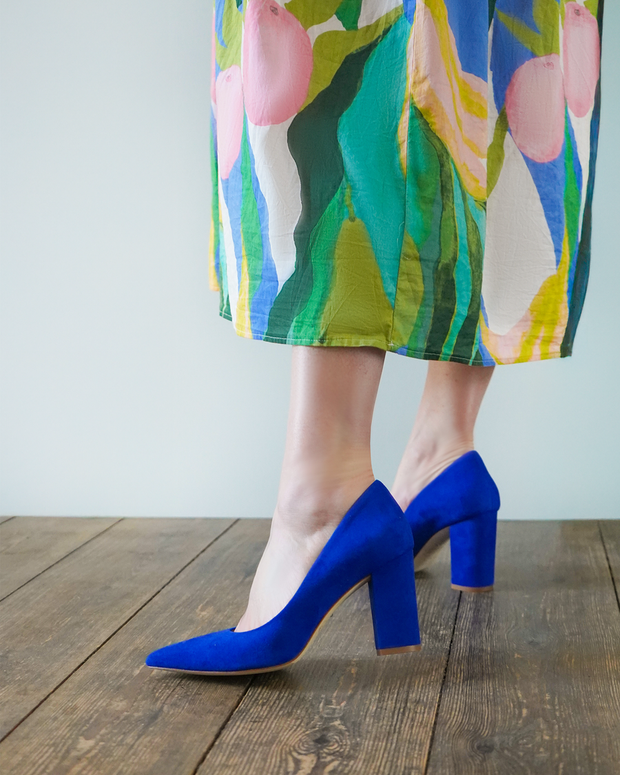 Blue suede heels with a block heel design, approximately 3.5 inches in height, worn by a person standing on a wooden floor. The background features a light-colored wall, and the individual is dressed in a colorful floral skirt that adds a vibrant contrast to the shoes. image