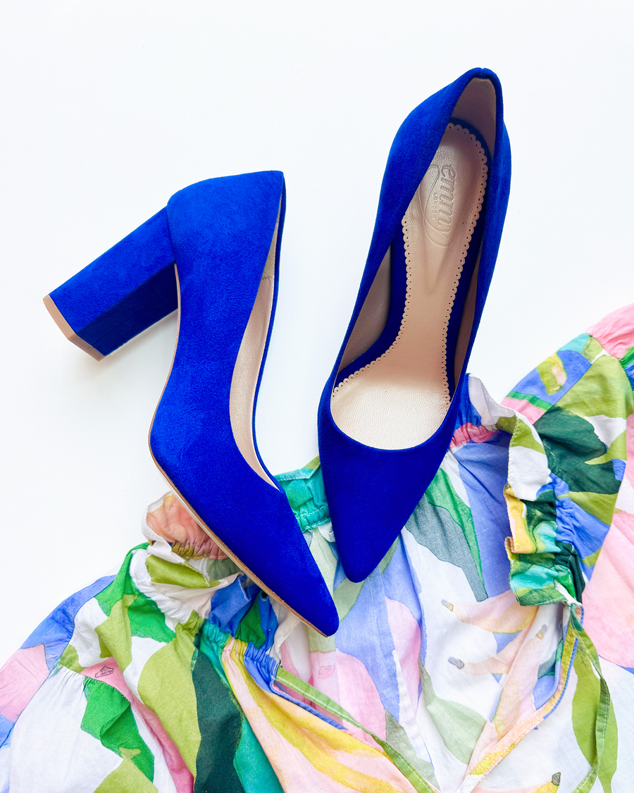 Blue suede high heels with a block heel, positioned on a colorful, patterned fabric. The shoes are displayed from a top-down angle, showcasing their pointed toe design and smooth interior. image