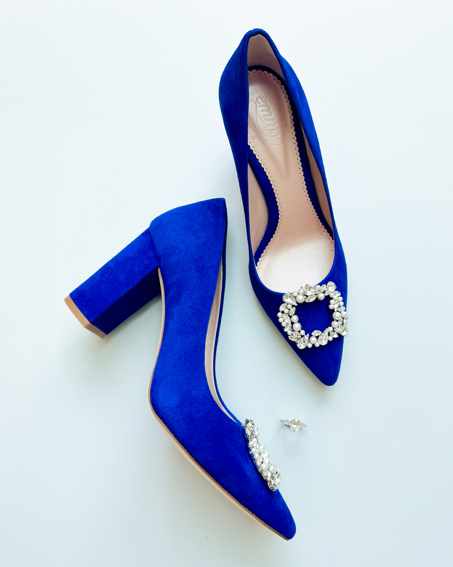 Blue suede heels with a chunky block heel, featuring a decorative rhinestone buckle on the toe. The shoes are positioned on a light background, showcasing their vibrant color and elegant design. image