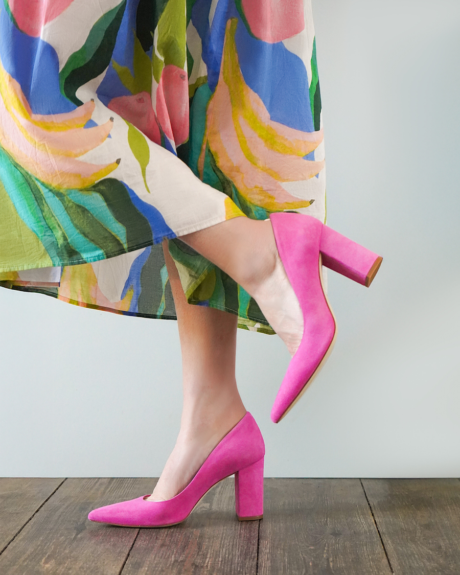 Pink suede heels with a block heel are worn by a person standing on a wooden floor. The background features a light-colored wall, and the individual is dressed in a colorful, floral-patterned skirt that flows around them. The angle of the image showcases the shoes from a side view, highlighting the heel and the overall design. image