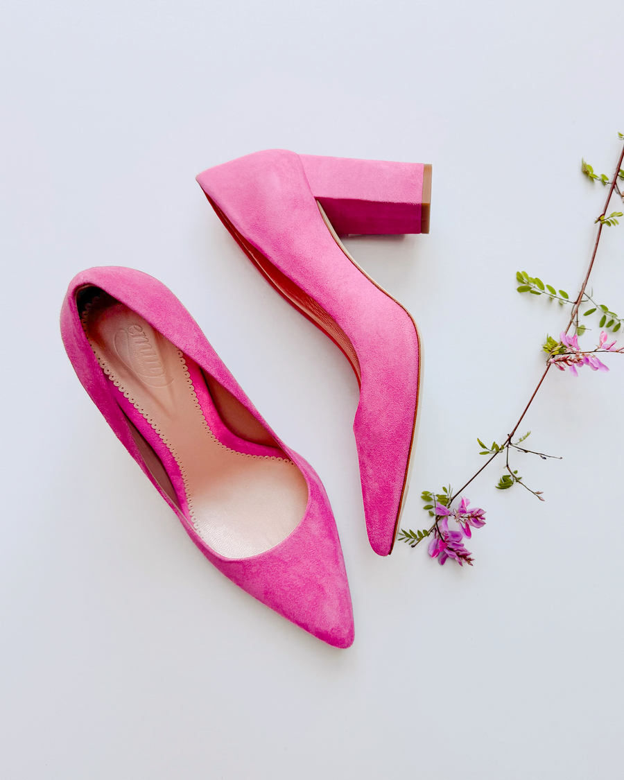 Pink suede heels with a chunky block heel, positioned on a white background. The shoes have a pointed toe and a sleek silhouette, surrounded by delicate pink flowers that add a touch of elegance to the composition. image