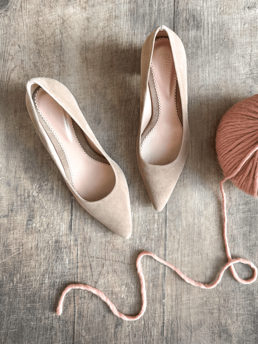 Nude suede pointed-toe heels displayed on a wooden surface, with a soft pink yarn ball and a strand of yarn nearby. The shoes are positioned from a top-down angle, showcasing their sleek design and elegant silhouette. image