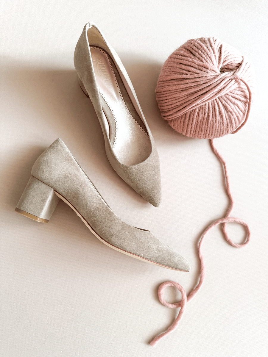 Beige suede heels with a block heel, positioned next to a pink yarn ball on a light background. The shoes have a pointed toe and are displayed from a slightly elevated angle, showcasing their elegant design. image