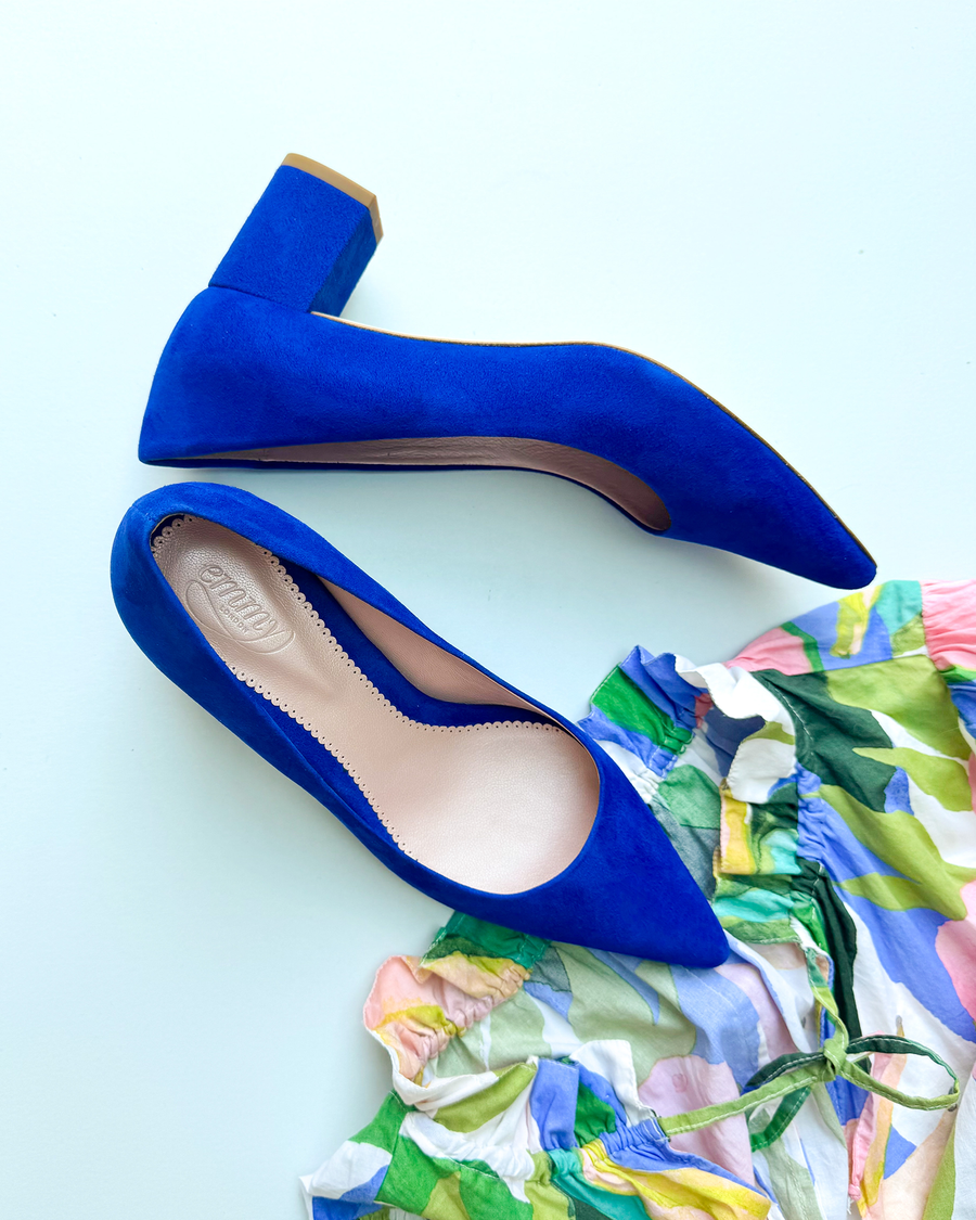 Blue suede heels with a block heel design are positioned on a light blue background. The shoes feature a pointed toe and are accompanied by a colorful, ruffled dress in the foreground, showcasing a vibrant mix of colors. image