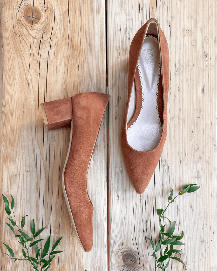 Brown suede heels with a block heel, positioned on a wooden surface with a natural grain. The shoes are pointed-toe style, showcasing a clean and minimalist design. Small green leaves are placed nearby, adding a touch of nature to the composition. image