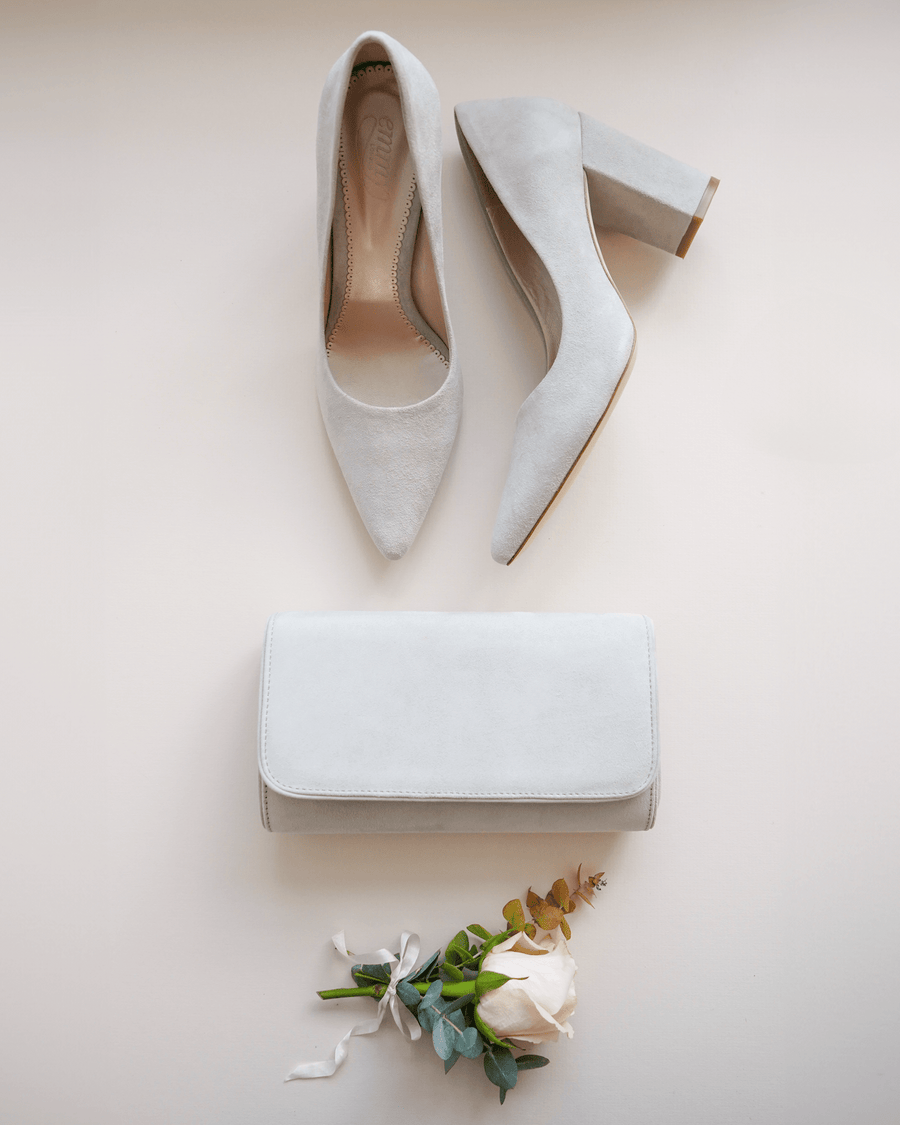 Elegant light grey suede heels with a block heel, positioned alongside a matching clutch and a delicate rose bouquet. The shoes are displayed from a top-down angle on a soft, neutral background, showcasing their pointed toe design. image