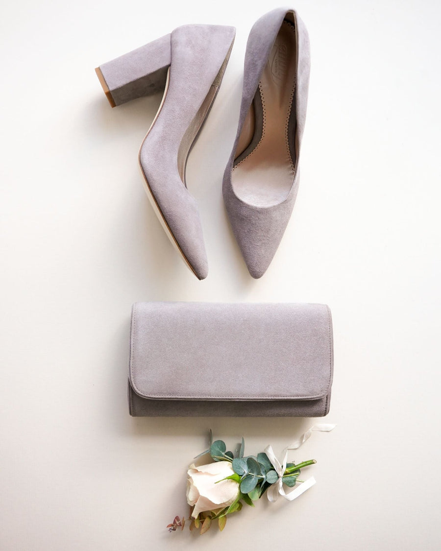 Gray suede high heels with a block heel are positioned above a matching gray clutch. The shoes are pointed-toe and are set against a light background, with a delicate arrangement of a white rose and greenery placed at the bottom of the image. The angle showcases the shoes from above, highlighting their sleek design. image