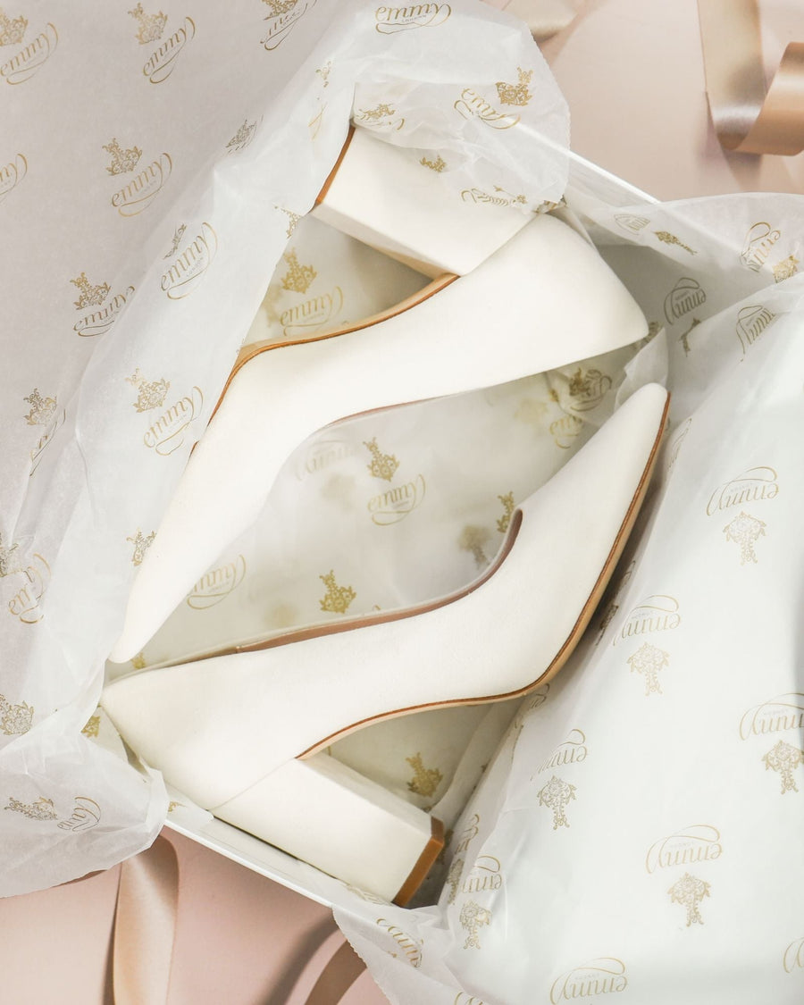 White leather heels with a block heel are displayed inside a gift box lined with tissue paper featuring a gold logo. The shoes are positioned at an angle, showcasing their pointed toe design. The background includes soft, neutral tones with a hint of ribbon, enhancing the elegant presentation. image