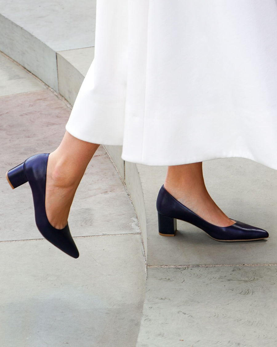 Elegant navy leather heels with a block heel, approximately 3 inches high, worn by a model standing on a smooth, light-colored surface. The model is dressed in a flowing white garment, and the angle captures the shoes from a side view, highlighting their sleek design. image