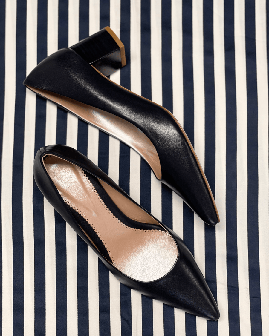 Elegant black leather heels with a pointed toe design, featuring a modest block heel. The shoes are positioned on a striped navy and white background, showcasing their sleek silhouette. The image captures the shoes from a top-down angle, highlighting the smooth finish and interior lining. image