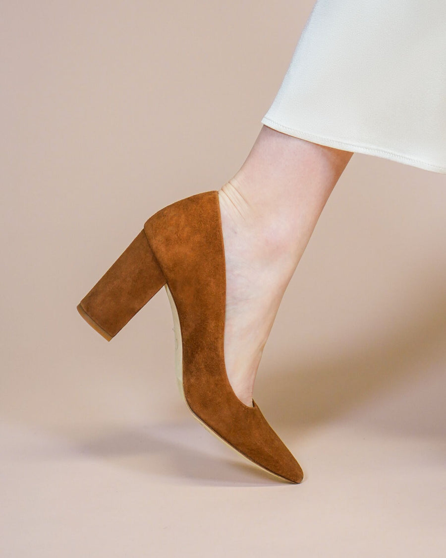 Brown suede high heel pump with a pointed toe, showcased at an angle against a soft beige background. The shoe is being worn, highlighting its sleek design and sturdy block heel. image