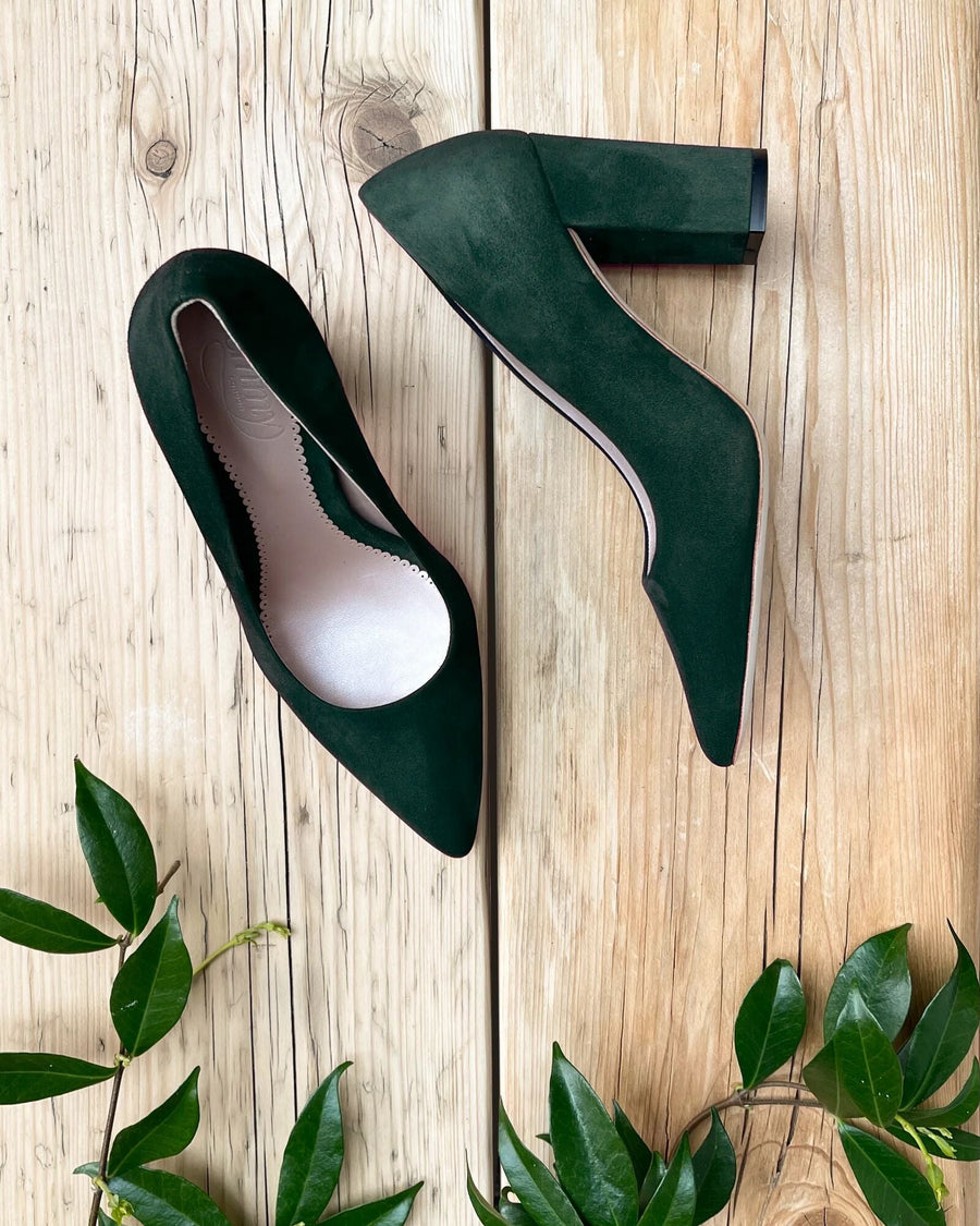 Green suede heels with a pointed toe and a chunky heel, positioned on a wooden surface surrounded by green leaves. The shoes are shown from a top-down angle, highlighting their sleek design and texture. image