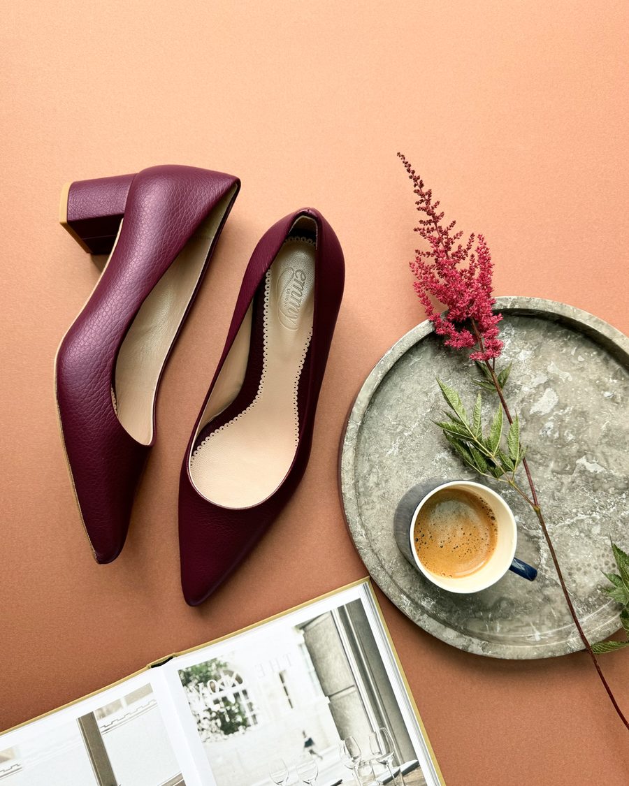 Elegant burgundy leather heels with a block heel design, positioned on a warm orange background. The shoes are displayed alongside a small cup of coffee and a decorative plate featuring greenery. A book is partially visible beneath the shoes, adding a touch of sophistication to the arrangement. image
