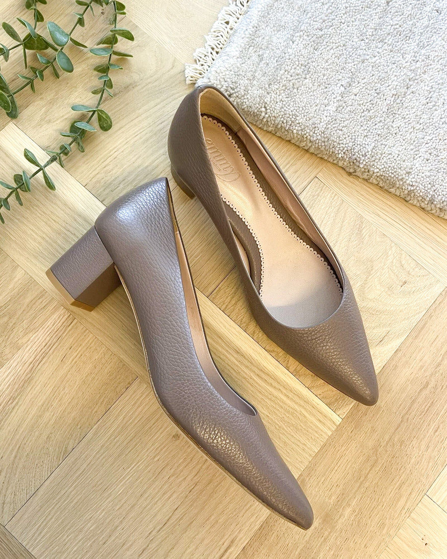 Gray leather heels with a block heel, approximately 3 inches high, are positioned on a light wooden floor. A textured beige rug is partially visible in the background, along with a small green plant, creating a warm and inviting atmosphere. The shoes are shown from a top-down angle, highlighting their pointed toe and elegant design. image