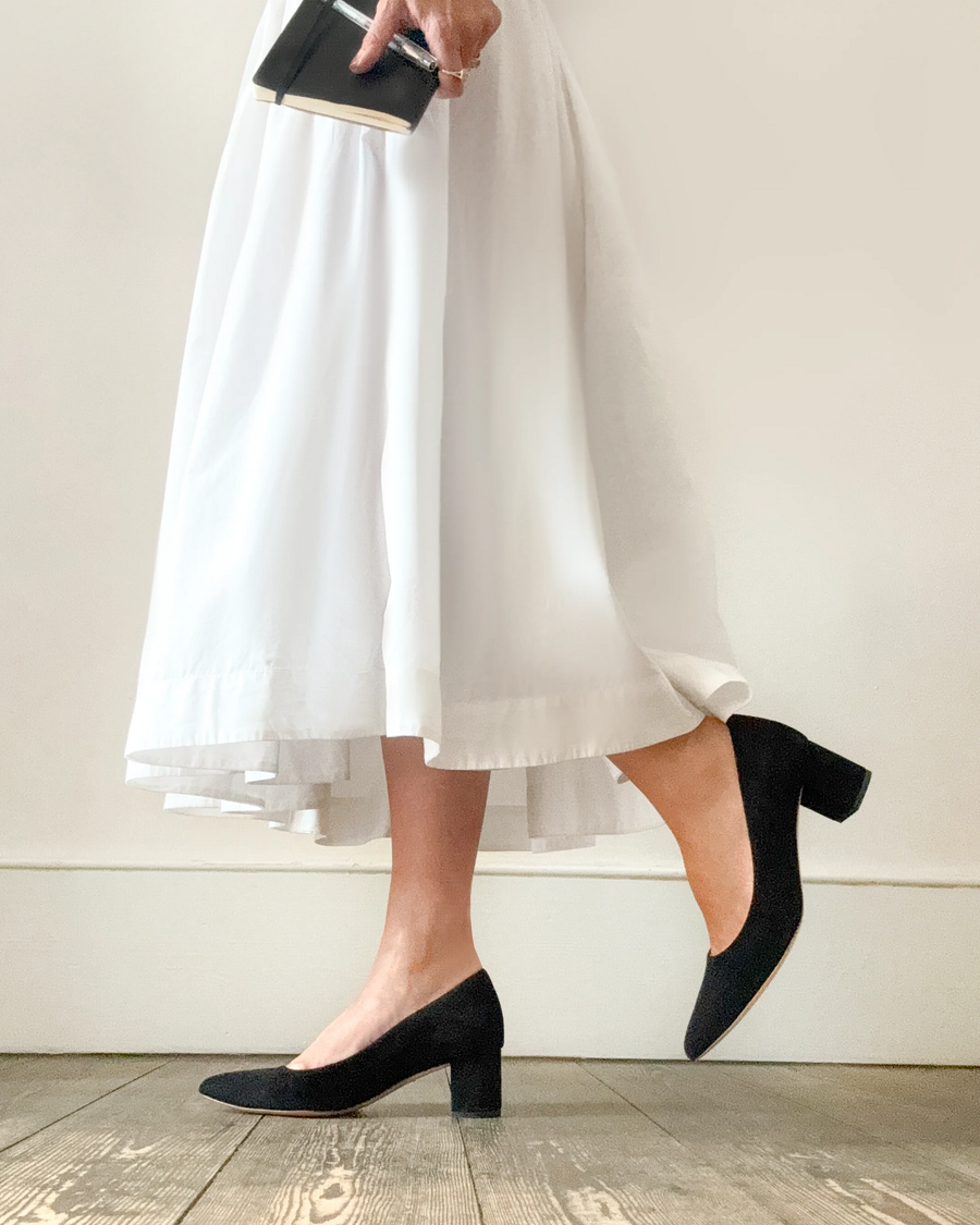 Black suede heels with a block heel are worn by a person in a white dress, standing on a wooden floor. The image captures the shoes from a side angle, showcasing the heel height and the elegant silhouette of the dress, with a neutral background that emphasizes the outfit. image