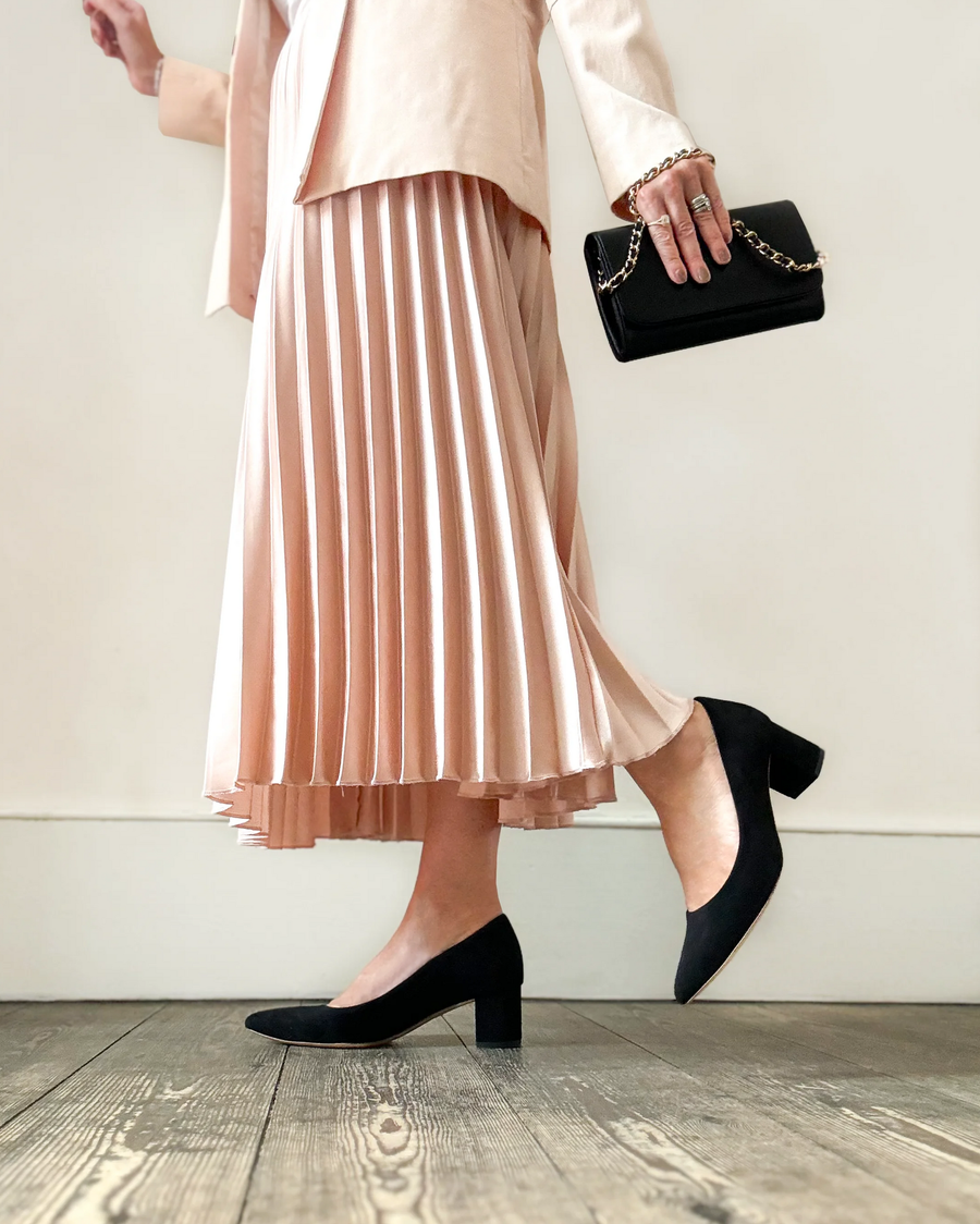 Black suede block heels are worn by a person in a light pink pleated skirt, standing on a wooden floor. The image is taken from a low angle, showcasing the shoes and the skirt's flow. The background is a plain white wall, emphasizing the outfit. image