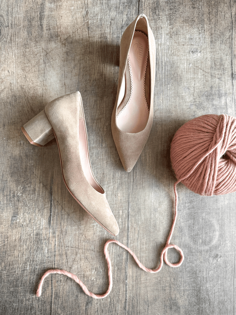 Beige suede heels with a block heel, positioned on a wooden surface. A pink yarn ball and a length of yarn are nearby, adding a cozy touch to the scene. The shoes are pointed-toe and are shown from a top-down angle, highlighting their elegant design. image