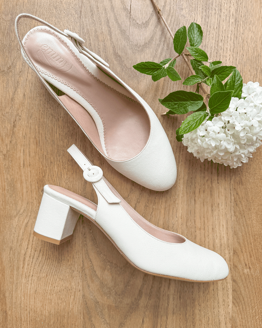 White slingback shoes with a square toe and a block heel, set against a wooden background. The shoes are made of leather and feature a delicate strap with a button closure. A sprig of green leaves and white flowers is placed nearby, adding a touch of nature to the scene. The angle showcases the shoes from above, highlighting their elegant design. image