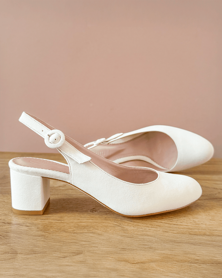 White suede slingback heels with a rounded toe and a chunky block heel. The shoes are positioned on a wooden surface against a soft pink background, showcasing their elegant design. image
