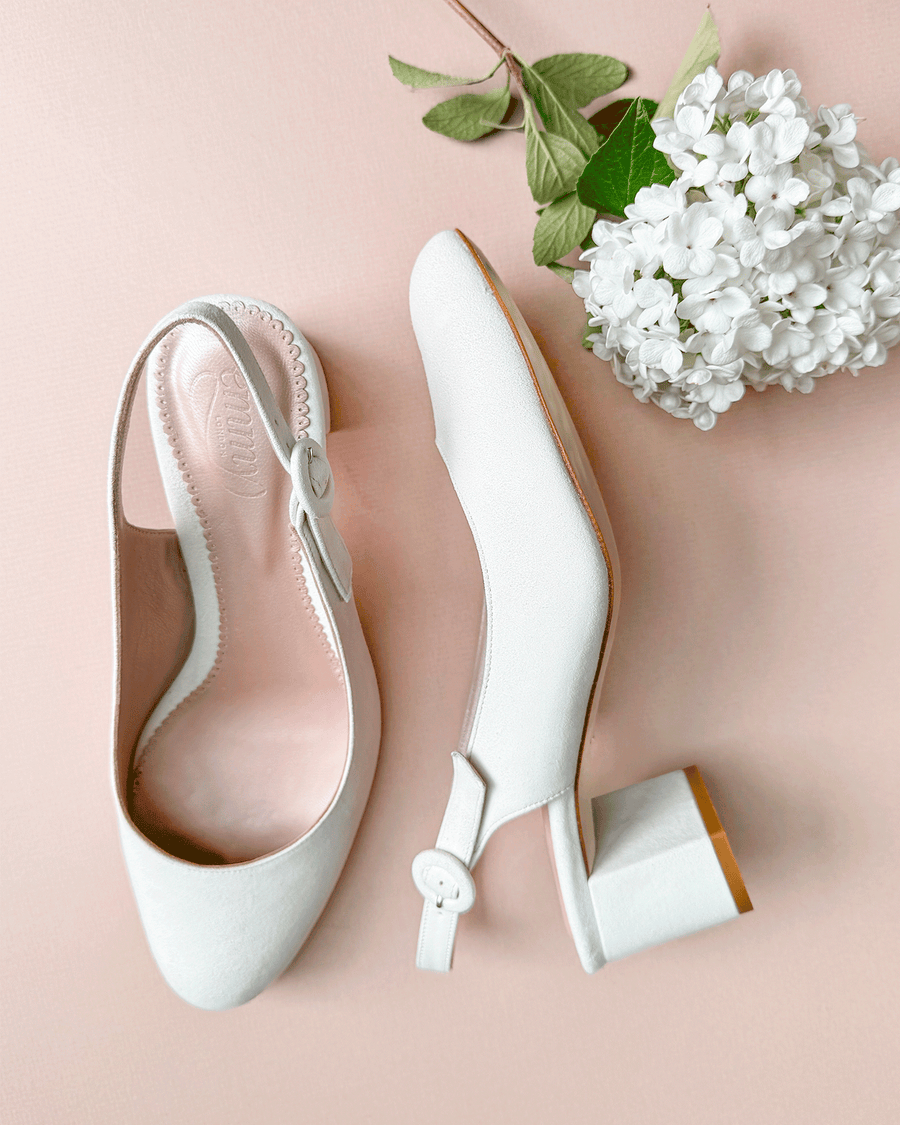 White leather slingback heels with a block heel, set against a soft pink background. The shoes feature a rounded toe and a delicate buckle detail on the side. A small bouquet of white flowers is positioned nearby, adding a touch of elegance to the composition. The angle showcases the shoes from above, highlighting their sleek design. image