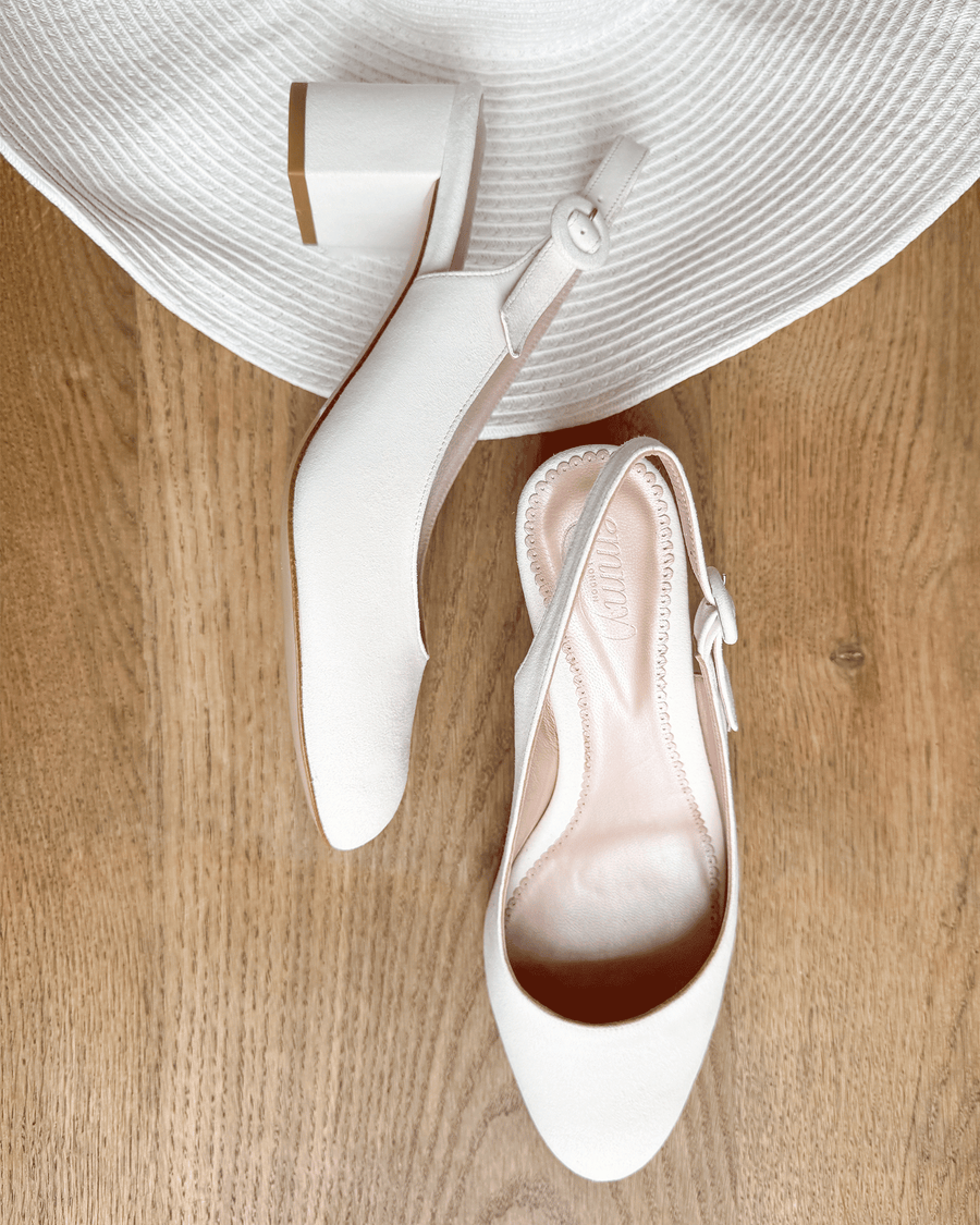 White slingback heels with a chunky block heel, set against a light circular straw mat on a wooden surface. The shoes feature a smooth finish and a delicate ankle strap, showcasing a clean and modern design. image