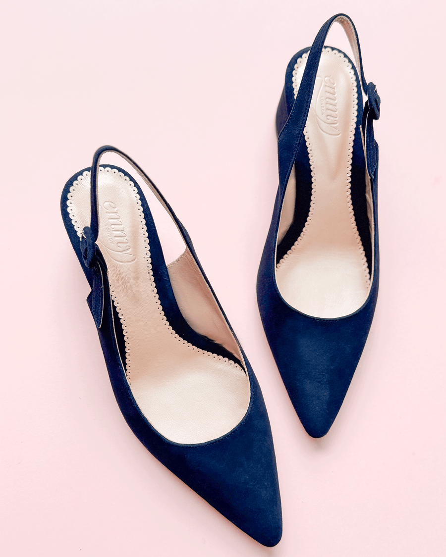Navy blue suede slingback heels with a pointed toe and a decorative bow on the side. The shoes are positioned on a light pink background, showcasing their elegant design. The angle captures the shoes from above, emphasizing their sleek silhouette. image