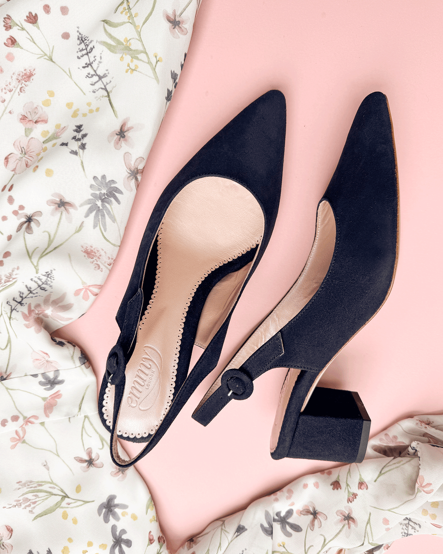 Black suede slingback heels with a block heel, featuring a pointed toe and a decorative button detail. The shoes are positioned on a soft pink background, surrounded by a floral-patterned fabric. The angle showcases the side view of the shoes, highlighting their elegant design. image