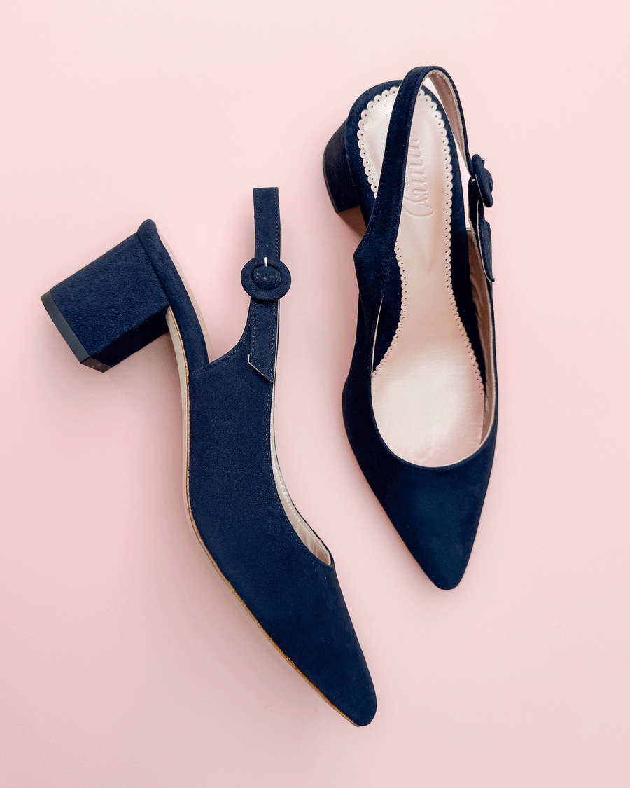 Navy blue suede slingback heels with a pointed toe and a chunky heel. The shoes are displayed on a soft pink background, showcasing their elegant design. The angle captures both the front and side view, highlighting the button detail on the strap. image