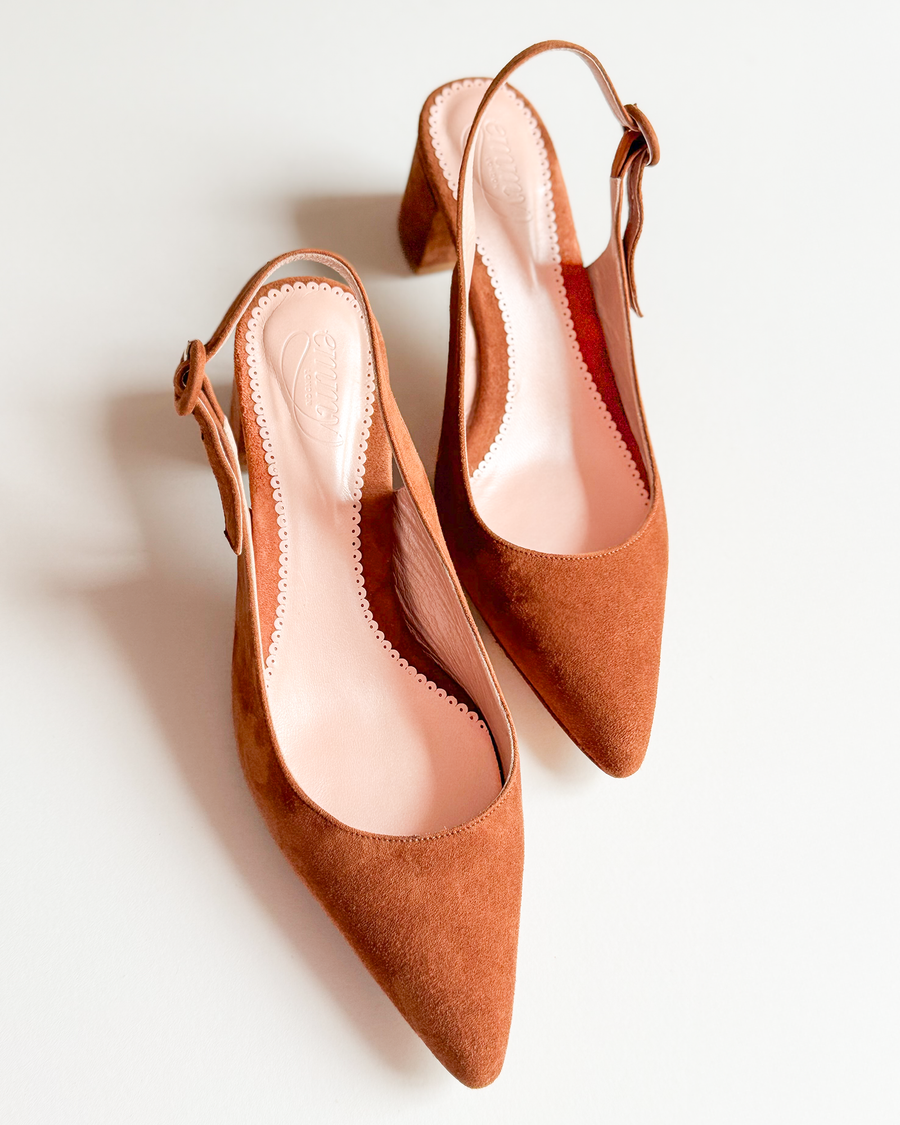 Brown suede slingback heels with a pointed toe design, set against a plain white background. The shoes feature a small block heel and a delicate bow detail on the side. The image captures the shoes from a top-down angle, showcasing their elegant silhouette. image