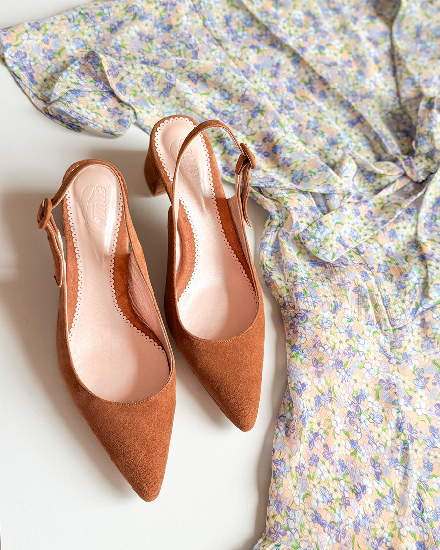 Slingback heels in a rich brown suede, featuring a pointed toe and a modest block heel. The shoes are positioned next to a light floral dress with a soft, flowing fabric, set against a neutral background. The angle captures the shoes from above, highlighting their elegant design and the texture of the suede. image