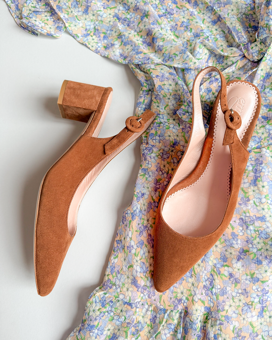 Brown suede slingback heels with a block heel, set against a floral-patterned dress. The shoes feature a rounded toe and a decorative button detail on the strap. The image is taken from a top-down angle, showcasing the shoes alongside the soft, pastel colors of the dress. image