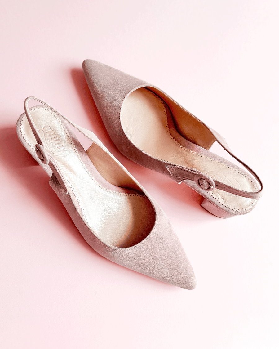 Pointed-toe slingback shoes in soft taupe suede, featuring a delicate button detail on the side. The shoes are positioned on a light pink background, showcasing their elegant design from a top-down angle. image