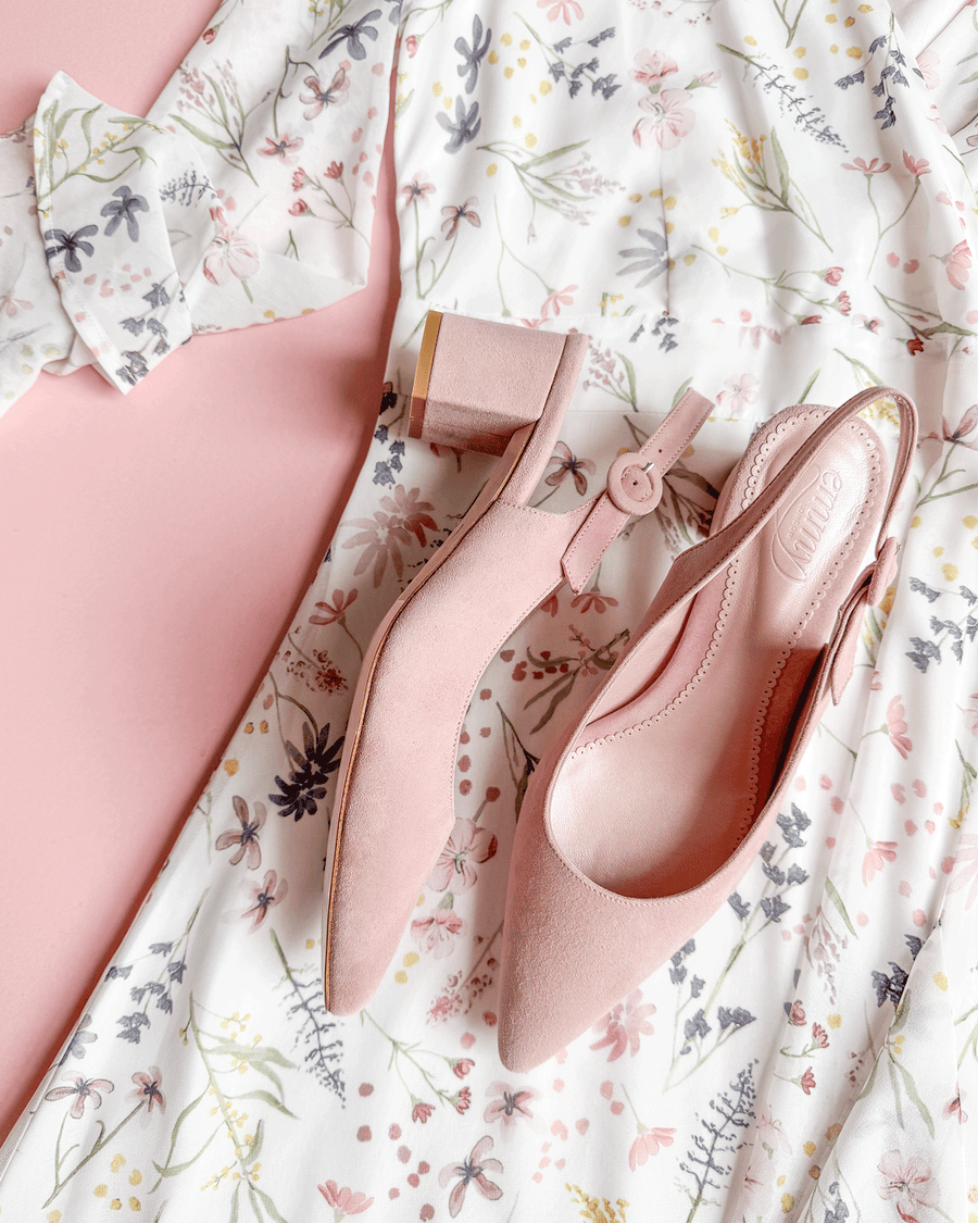 Pink suede slingback heels are positioned on a floral-patterned dress with pastel colors. The shoes feature a block heel and an ankle strap, showcasing a side view. The background is a soft pink, enhancing the delicate aesthetic of the ensemble. image